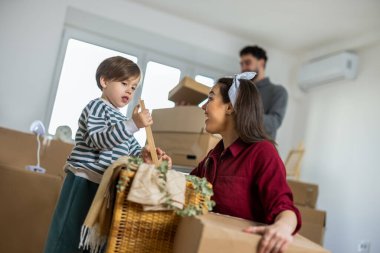 Yeni bir dairede, bir annenin bebeğiyle oynamak için diz çöktüğü ve onu güldürdüğü şefkatli bir aile anı. Bulanık arkaplanda baba kutuları düzenleyip açmakla meşgul görünüyor..