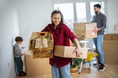 Yeni dairesinde, elinde küçük bir karton kutuyla kameraya gülümseyen genç bir annenin canlı bir portresi. Bulanık arka planda, kocası ve küçük oğlu görülebiliyor ve aile kavgasına içerik katıyor.
