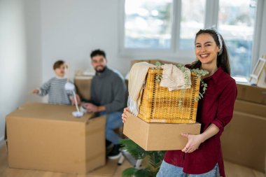 Yeni dairesinde, elinde küçük bir karton kutuyla kameraya gülümseyen genç bir annenin canlı bir portresi. Bulanık arka planda, kocası ve küçük oğlu görülebiliyor ve aile kavgasına içerik katıyor.