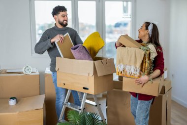 Genç bir erkek ve kadın arasında taşınma sırasında romantik ve güçlü bir an. İkisinin de elinde karton kutular var, ama gözleri sevgi dolu bir bakışla kilitlenmiş, başarının ve ortak hayallerin simgesi olan bir gülümsemeyi paylaşıyorlar..