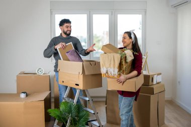 Genç bir erkek ve kadın arasında taşınma sırasında romantik ve güçlü bir an. İkisinin de elinde karton kutular var, ama gözleri sevgi dolu bir bakışla kilitlenmiş, başarının ve ortak hayallerin simgesi olan bir gülümsemeyi paylaşıyorlar..
