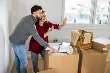 Karton kutuların arasında dikilen bir adam ve kadın duvara doğru hareket ediyor ve yeni evleri için iç tasarım fikirlerini tartışıyorlar..