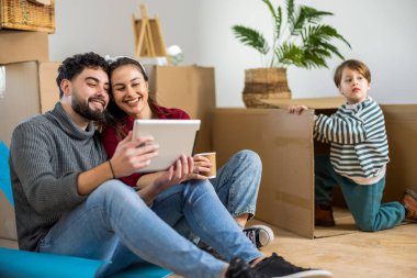 Genç bir çift boş evlerinin zemininde oturuyor, tablet kullanıyor ve paket kahve içiyorlar, küçük oğulları arka planda kutuları taşırken.