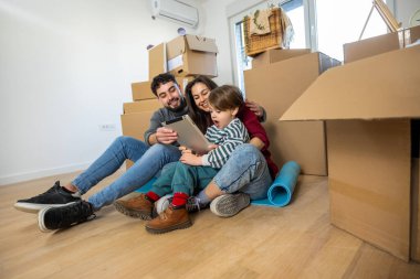 Neşeli bir anne ve baba yerde oturup tablet ekranda eğitimsel bir oyun izliyorlar. Meraklı çocukları boş bir dairede..