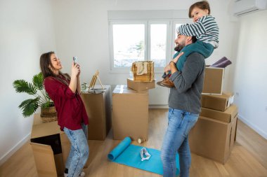 Neşeli bir baba oğlunu omuzlarında taşırken anne mutlu anılarını telefon kamerasıyla yakalıyor. Paketlenmemiş eşyalarla çevrili bir ailenin heyecanını, bağlantısını ve neşesini sembolize ediyor..