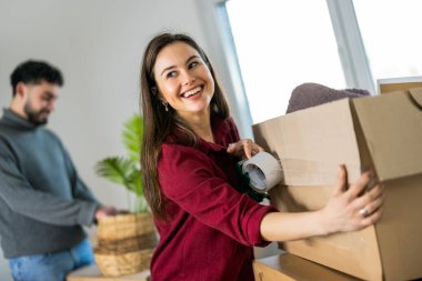 Odaklanmış ve mutlu bir kadın, yeni evindeki taşınma kutusundan bir şeyler çıkarır. Ortağı da arkasında bulanıklaşır. Yerleştirme ve yerel takım çalışmalarının yoğun atmosferini yakalamak.