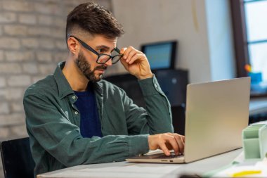 Modern bir ofisteki bir göreve odaklanmış ciddi bir profesyonel. Yoğun bir iş yükünü yönetirken genç bir adamın taşınabilir bilgisayar kullanırken gizli bir fotoğrafı..