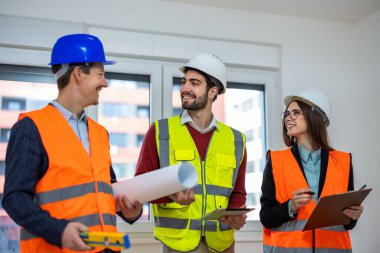 A professional construction crew in hard hats and vests having a discussion. They are talking and looking at each other while planning a renovation in an empty room.