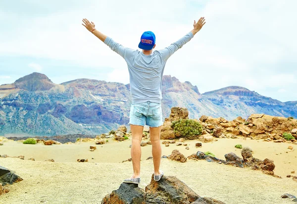 Happy stylish man in casual hipster clothes standing on the cliff of mountain with raised hands to the sun and celebrating success — Stock Photo, Image