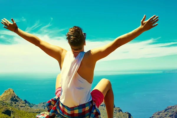 Feliz hombre elegante en ropa hipster casual de pie en el acantilado de la montaña con las manos levantadas al sol y celebrando el éxito —  Fotos de Stock