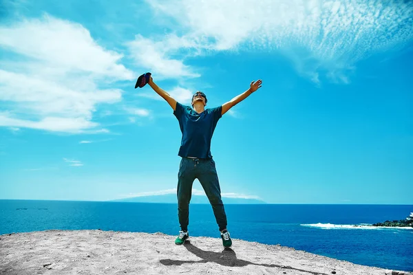 Happy stylish man in casual clothes standing on the cliff of mountain with raised hands to the sun and celebrating success — Stock Photo, Image