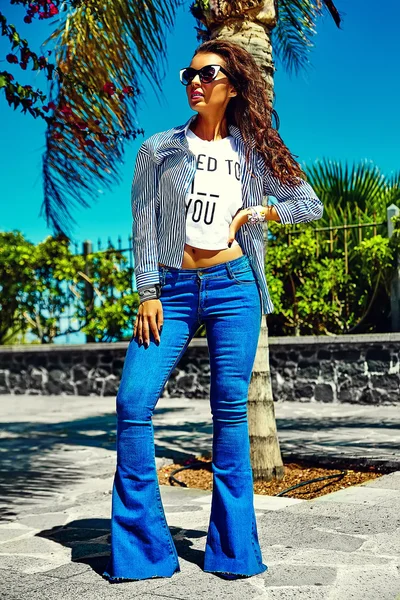 Fashion stylish beautiful young smiling brunette woman model in summer hipster casual blue flared jeans clothes posing in the street — Stock Photo, Image