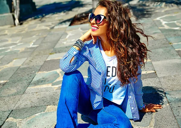 Модная красивая молодая улыбающаяся брюнетка модель в летней хипстерской повседневной синей одежде, сидящая на улице в солнцезащитных очках — стоковое фото