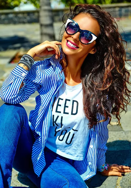 Fashion stylish beautiful young smiling brunette woman model in summer hipster casual blue clothes sitting in the street in sunglasses — Stock Photo, Image