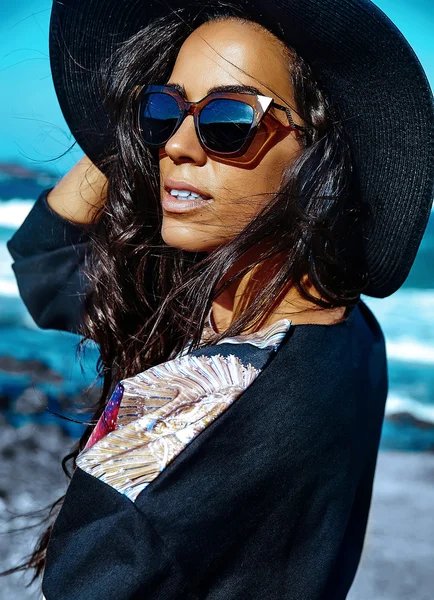 Fashion stylish beautiful young smiling brunette woman model in summer hipster casual clothes posing behind blue ocean and sky background in hat and sunglassesfashion stylish beautiful young smiling brunette woman model in summer hipster casual cloth — Stock Photo, Image