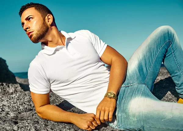 Young stylish handsome man model in hipster summer clothes posing on beach behind sky and ocean — Stock Photo, Image