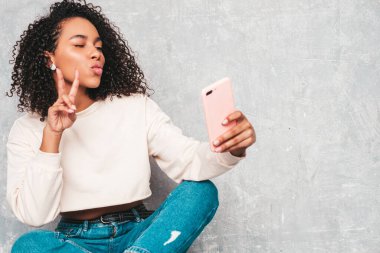 Saçları kıvırcık, güzel siyah bir kadın. Beyaz renkli süveter içinde gülümseyen bir model. Stüdyoda gri duvarın yanında oturan seksi, kaygısız kadın. Selfie çekerek hava öpücüğü veriyor.