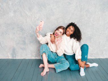 İki güzel, güzel, gülümseyen uluslararası hippi kadın, son moda yaz kot kıyafetleri içinde. Stüdyoda gri duvarın yanında oturan kaygısız seksi kadınlar. Pozitif modeller eğleniyor. Selfie çekiyorlar.
