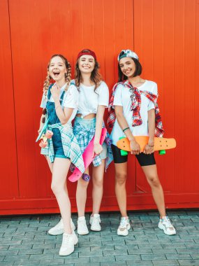 Renkli paten kaykayan üç genç ve güzel bayan. Yazlık hippi elbiseli kadınlar kırmızı duvarın yanında poz veriyor. Pozitif mankenler eğleniyor ve deliriyor.