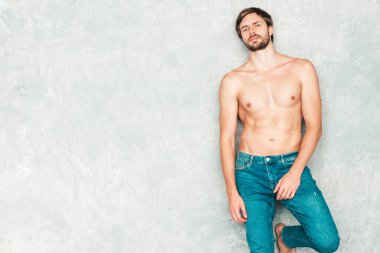 Yakışıklı, güçlü bir adamın portresi. Kot pantolonla gri duvarın yanında poz veren sağlıklı gülümseyen atletik form modeli. Çıplak gövdesi olan kendine güvenen seksi moda erkeği. Lambersexual stüdyoda.