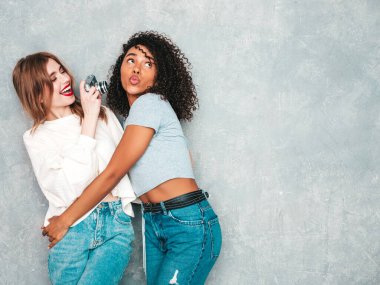İki güzel, güzel, gülümseyen uluslararası hippi kadın, son moda yaz kot kıyafetleri içinde. Stüdyoda gri duvarın yanında poz veren kaygısız seksi kadınlar. Pozitif modeller eğleniyor. Retro kamerada fotoğraf çekmek