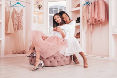 Son moda yaz elbiseleri içinde gülümseyen iki güzel uluslararası hippi kadın. Pembe gardıropta poz veren kaygısız seksi kadınlar. Pozitif modeller eğleniyor. Kanepede oturuyorlar.