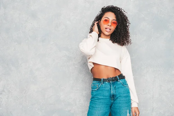 Beautiful black woman with afro curls hairstyle.Smiling model in white trendy jeans clothes and sunglasses. Sexy carefree female posing near gray wall in studio. Tanned and cheerful