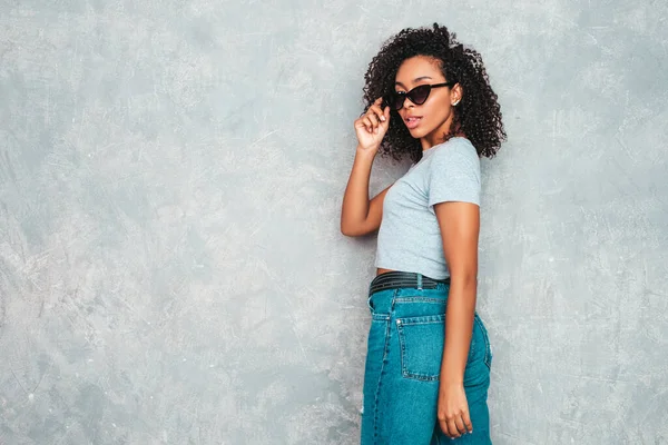 Beautiful black woman with afro curls hairstyle.Smiling model in trendy jeans clothes and sunglasses. Sexy carefree female posing near gray wall in studio. Tanned and cheerful