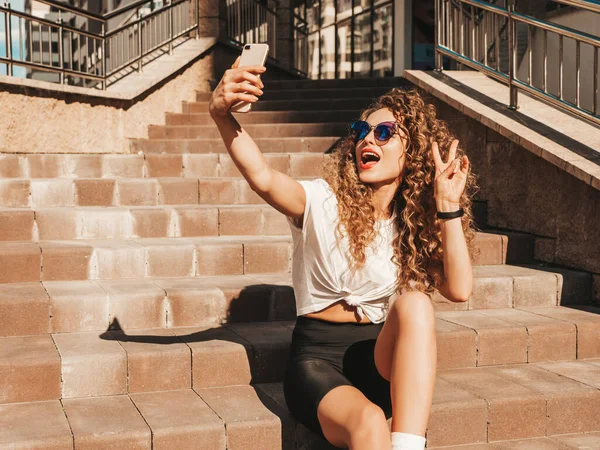 Beautiful smiling model with afro curls hairstyle dressed in summer hipster clothes.Sexy carefree female sitting on  stairs in the street in sunglasses.Taking selfie self portrait photos on smartphone