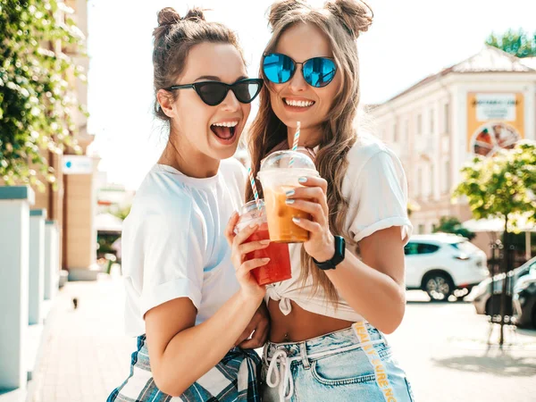 流行の夏服の2人の若い美しい笑顔ヒップスター女性 屋外でポーズをとる気の利いた女性 積極的なモデルを保持し わらとプラスチックカップで新鮮なカクテルスムージードリンクを飲みます — ストック写真