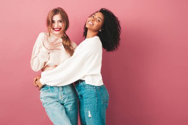 İki güzel, güzel, gülümseyen uluslararası hippi kadın, son moda yaz kot kıyafetleri içinde. Stüdyoda pembe duvarın yanında poz veren kaygısız seksi kadınlar. Pozitif modeller eğleniyor. Arkadaşlık kavramı 