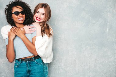 İki güzel, güzel, gülümseyen uluslararası hippi kadın, son moda yaz kot kıyafetleri içinde. Stüdyoda gri duvarın yanında poz veren kaygısız seksi kadınlar. Pozitif modeller eğleniyor. Arkadaşlık kavramı 