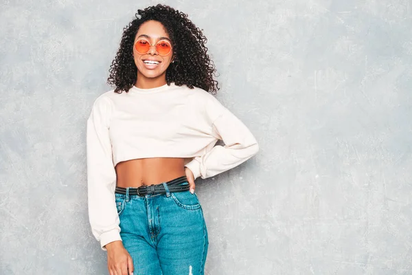 Beautiful black woman with afro curls hairstyle.Smiling model in  trendy jeans clothes and sunglasses. Sexy carefree female posing near gray wall in studio. Tanned and cheerful