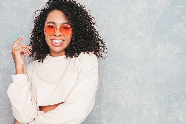 Beautiful black woman with afro curls hairstyle.Smiling model in  trendy jeans clothes and sunglasses. Sexy carefree female posing near gray wall in studio. Tanned and cheerful