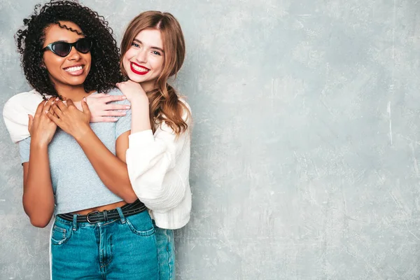 İki güzel, güzel, gülümseyen uluslararası hippi kadın, son moda yaz kot kıyafetleri içinde. Stüdyoda gri duvarın yanında poz veren kaygısız seksi kadınlar. Pozitif modeller eğleniyor. Arkadaşlık kavramı 