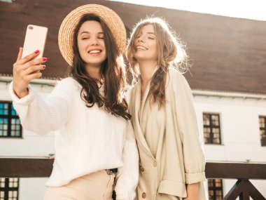 Son moda beyaz süveter ve palto giymiş iki güzel, gülümseyen hippi kadın. Sokakta şapkayla poz veren seksi kaygısız kadınlar. Pozitif modeller dışarıda eğleniyor. Fotoğraf çekiyorum.