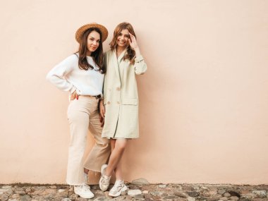 Son moda beyaz süveter ve palto giymiş iki güzel, gülümseyen hippi kadın. Sokakta arka planda duvarın yanında şapkayla poz veren kaygısız seksi kadınlar. Pozitif modeller dışarıda eğleniyor.