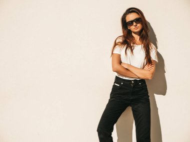 Güneş gözlüklü kendine güvenen güzel bir modelin portresi. Yazlık hippi tişörtü ve kot pantolon giymiş bir kadın. Sokakta duvar kenarında poz veriyor. Çapraz kollu kadın