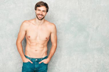 Yakışıklı, güçlü bir adamın portresi. Kot pantolonla gri duvarın yanında poz veren sağlıklı gülümseyen atletik form modeli. Çıplak gövdesi olan kendine güvenen seksi moda erkeği. Lambersexual stüdyoda.
