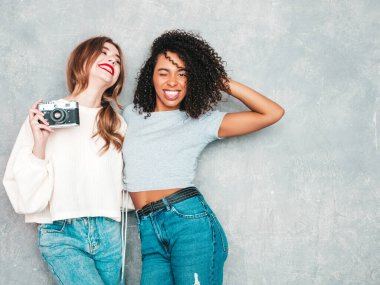 İki güzel, güzel, gülümseyen uluslararası hippi kadın, son moda yaz kot kıyafetleri içinde. Stüdyoda gri duvarın yanında poz veren kaygısız seksi kadınlar. Pozitif modeller eğleniyor. Retro kamerada fotoğraf çekmek