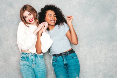 İki güzel, güzel, gülümseyen uluslararası hippi kadın, son moda yaz kot kıyafetleri içinde. Stüdyoda gri duvarın yanında poz veren kaygısız seksi kadınlar. Pozitif modeller eğleniyor. Arkadaşlık kavramı 