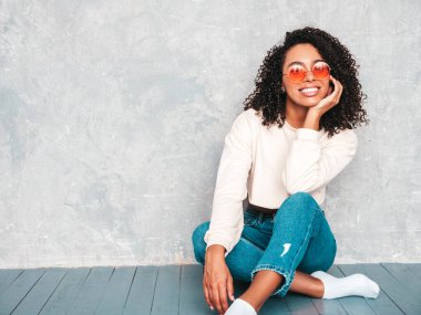 Saçları kıvırcık, güzel siyah bir kadın. Moda kotlar ve güneş gözlükleriyle gülümseyen bir model. Stüdyoda gri duvarın yanında oturan kaygısız seksi kadın.