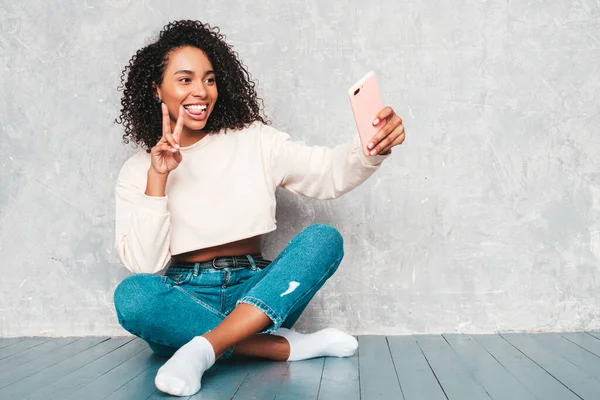 Beautiful black woman with afro curls hairstyle.Smiling model in trendy jeans clothes and sunglasses. Sexy carefree female sitting on the floor near gray wall in studio.She taking selfie photos