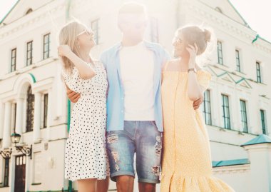 Sokakta poz veren üç genç arkadaş grubu. Moda adamı ve sıradan yaz kıyafetleri giymiş iki tatlı kadın. Gülümseyen modeller güneş gözlükleriyle eğleniyorlar. Neşeli kadınlar ve gün batımındaki adam.