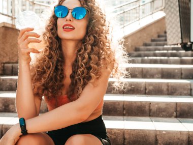 Saçları kıvırcık, genç ve güzel bir hippi kadın. Yazın modaya uygun giyinmiş, sokakta poz veren bir kadın. Modeli elinde tutuyor ve samanla plastik bardakta taze kokteyl içiyor.