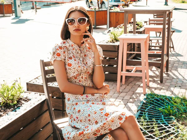 Portrait of young beautiful smiling  woman in trendy summer sundress.Sexy carefree woman sitting in veranda cafe on the street background at sunset. Positive model outdoors in sunglasses