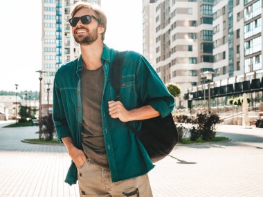 Yakışıklı, gülümseyen stil sahibi Lambersex modelinin portresi. Yeşil tişörtlü adam. Moda erkek gökdelen arka planında güneş gözlükleriyle sokakta poz veriyor. Gün batımında sırt çantasıyla dışarıda.