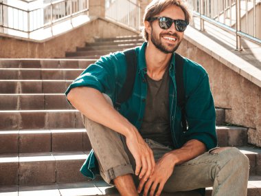 Yakışıklı, gülümseyen stil sahibi Lambersex modelinin portresi. Yeşil tişörtlü adam. Moda erkeği, güneş gözlüklü arka planda, merdivenlerde oturuyor.
