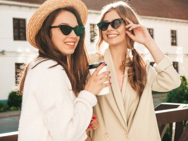 Son moda beyaz süveter ve palto giymiş iki güzel, gülümseyen hippi kadın. Sokakta şapka ve güneş gözlükleriyle poz veren kaygısız seksi kadınlar. Pozitif modeller eğleniyor ve kahve içiyor. 