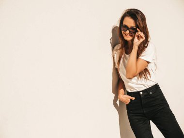 Güneş gözlüklü güzel gülümseyen modelin portresi. Yazlık hippi tişörtü ve kot pantolon giymiş bir kadın. Sokakta duvar kenarında poz veriyor. Komik ve pozitif kadın dışarıda eğleniyor.